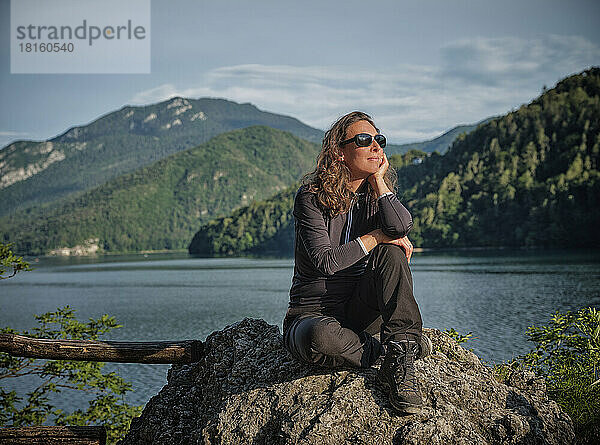 Lächelnde  nachdenkliche Frau sitzt auf einem Felsen vor dem See