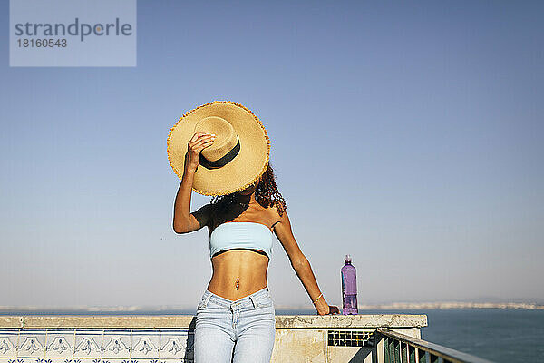 Junge Frau bedeckt Gesicht mit Hut und steht an sonnigem Tag an der Wand