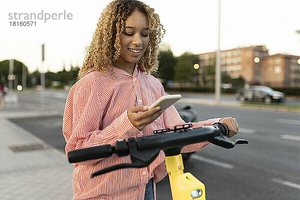 Lächelnde Frau mit Elektroroller und Mobiltelefon auf Fußweg