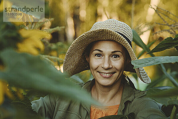 Lächelnde Frau mit Hut inmitten grüner Blätter im Garten
