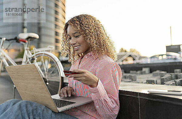 Lächelnde Frau mit Kreditkarte beim Online-Shopping am Laptop neben der Bank