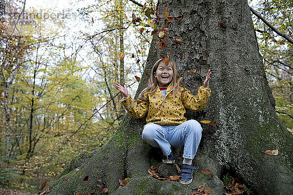 Fröhliches Mädchen  das mit Blättern spielt und auf einem Baumstamm im Wald sitzt