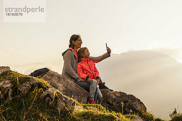 Glückliche Mutter mit Tochter  die auf einem Felsen sitzt und ein Selfie mit dem Handy macht