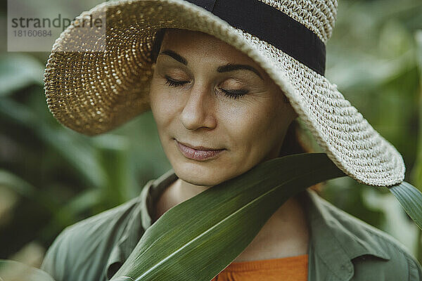 Reife Frau mit geschlossenen Augen berührt grünes Blatt im Gesicht im Garten
