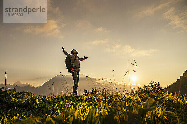 Reifer Mann steht bei Sonnenuntergang mit ausgestreckten Armen auf dem Gipfel des Berges