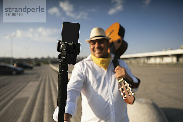 Lächelnder Gitarrist hält Gitarre und macht ein Selfie mit dem Mobiltelefon auf einem Stativ