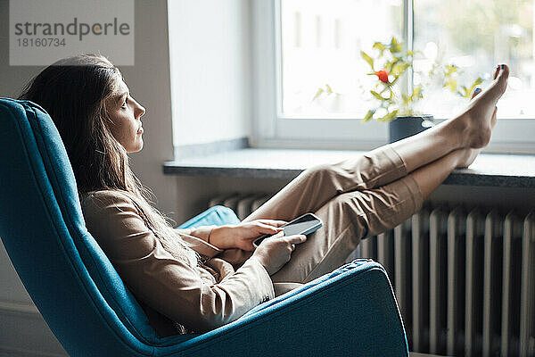 Nachdenkliche junge Geschäftsfrau sitzt zu Hause mit erhobenen Füßen auf einem Stuhl