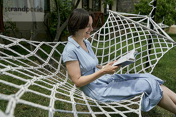 Lächelnde Frau liest ein Buch und sitzt auf einer Hängematte im Garten