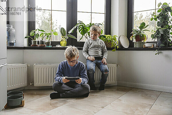 Junge sitzt mit Bruder am Heizkörper und benutzt zu Hause Smartphone