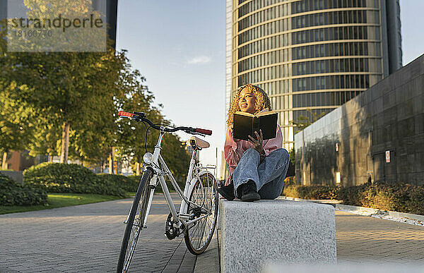 Lächelnde  nachdenkliche Frau mit Buch  die mit dem Fahrrad auf einer Bank sitzt