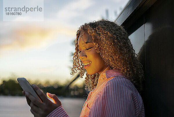Junge Frau mit lockigem Haar benutzt Smartphone bei Sonnenuntergang