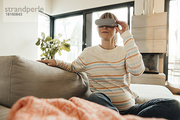 Werdende Frau mit Virtual-Reality-Headset sitzt zu Hause auf dem Sofa