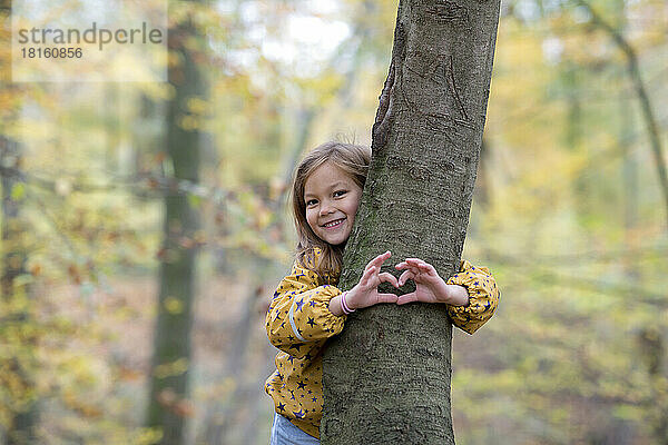 Lächelndes süßes Mädchen umarmt Baum und gestikuliert in Herzform im Wald