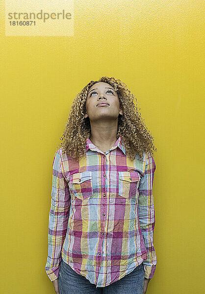 Nachdenkliche junge Frau blickt vor gelbem Hintergrund nach oben