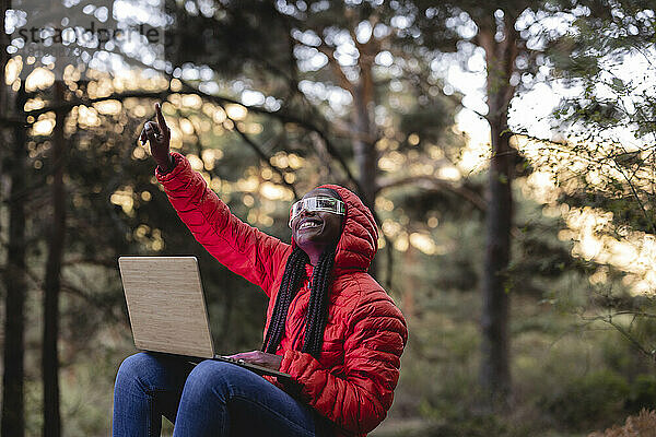 Glückliche Frau mit futuristischer Brille und Laptop gestikuliert im Wald