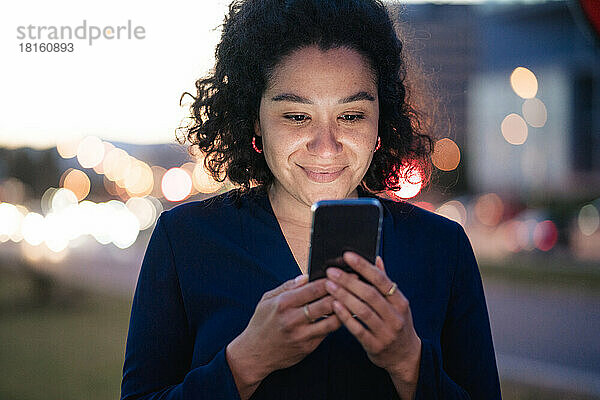 Lächelnde Geschäftsfrau  die in der Abenddämmerung ihr Mobiltelefon benutzt