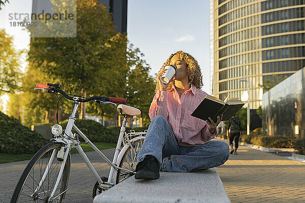 Junge Frau mit Buch trinkt Kaffee auf Bank in der Stadt