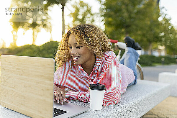 Lächelnde junge Frau mit Einwegkaffeetasse und Laptop auf der Bank