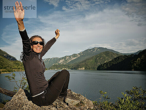 Glückliche Frau mit erhobenen Armen auf einem Felsen vor dem See