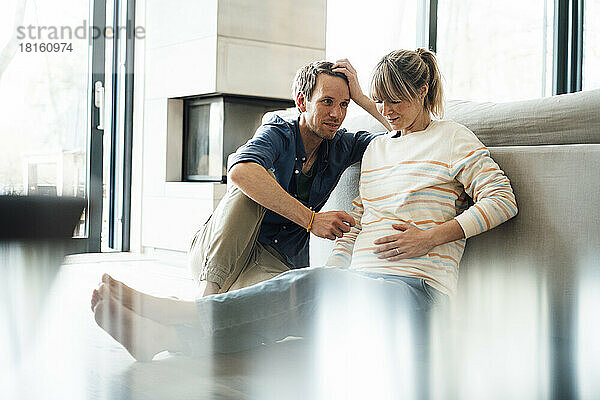 Mann spricht mit schwangerer Frau  die zu Hause sitzt und ihren Bauch berührt