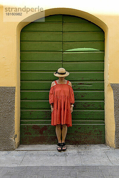 Ältere Frau in rotem Kleid und Sonnenhut vor grüner Tür