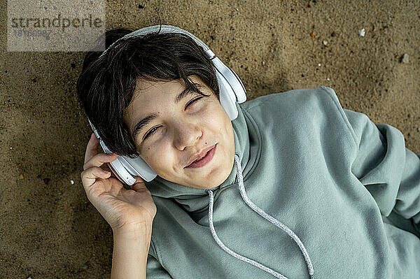 Lächelnder Junge mit kabellosen Kopfhörern  der Musik hört und sich im Sand entspannt