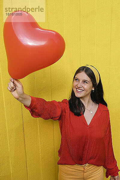 Glückliche Frau hält herzförmigen Ballon vor gelber Wand