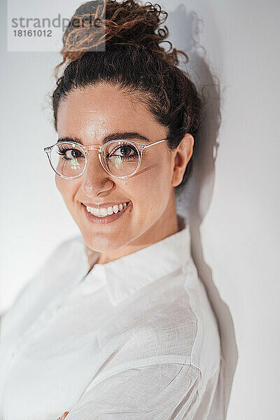 Lächelnde Frau mit Brille lehnt zu Hause an weißer Wand