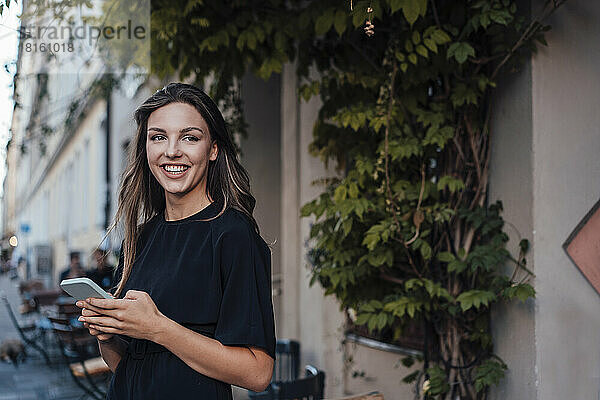 Fröhliche junge Frau mit Smartphone steht neben Pflanzen