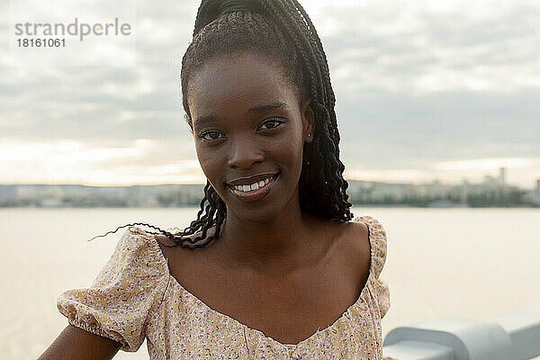 Lächelnde junge Frau steht am Geländer vor dem Fluss