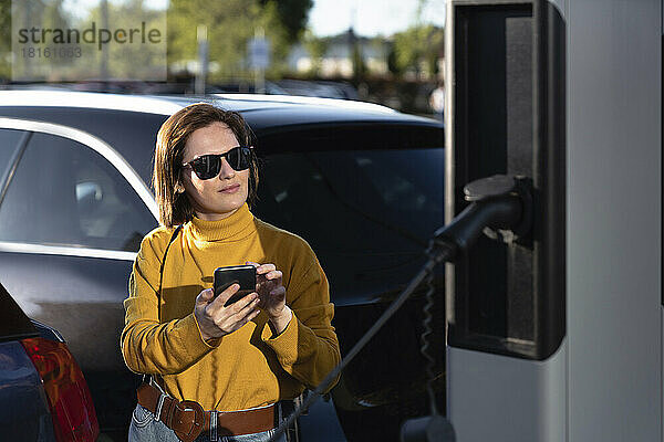 Frau mit Sonnenbrille steht mit Mobiltelefon an Fahrzeugladestation