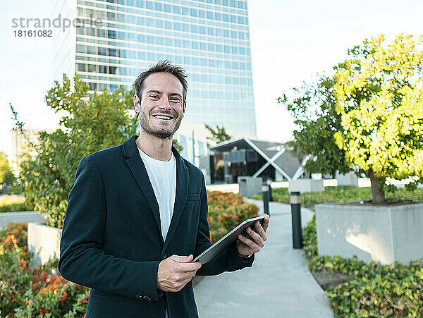 Glücklicher Geschäftsmann mit Tablet-PC steht auf Fußweg im Büropark