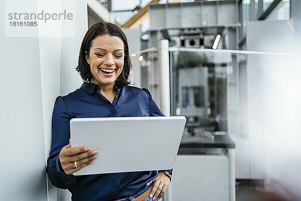 Glückliche Geschäftsfrau blickt auf Tablet-PC  der in der Industrie an der Wand lehnt