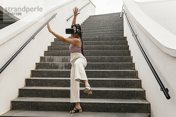 Junge Frau mit Virtual-Reality-Simulator steht mit erhobener Hand auf der Treppe