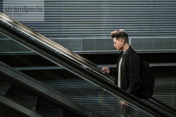 Junger Mann benutzt Smartphone auf Rolltreppe