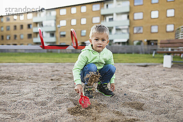 Netter Junge spritzt Sand mit Schaufel auf dem Spielplatz