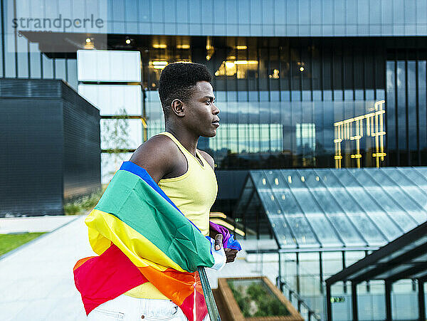 Selbstbewusster junger Mann  gehüllt in eine Regenbogenfahne  lehnt am Geländer moderner Glasgebäude