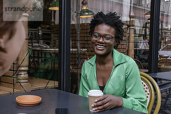 Glückliche Frau hält Einwegbecher in der Hand und unterhält sich mit einer Freundin im Straßencafé