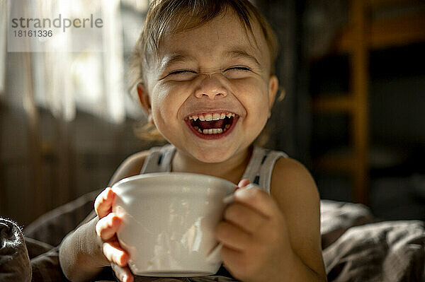 Cute baby with cup laughing in bed at home