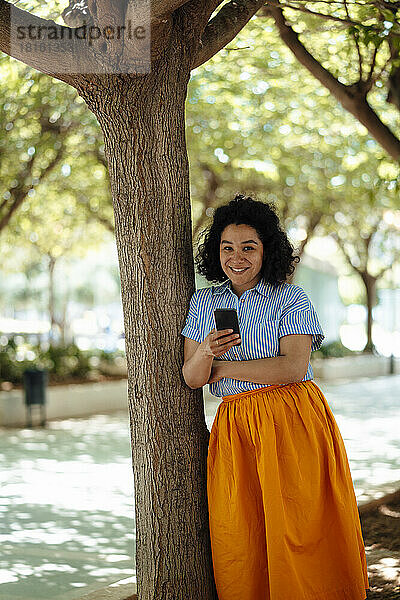 Lächelnde Frau mit Mobiltelefon  die sich an einen Baumstamm lehnt