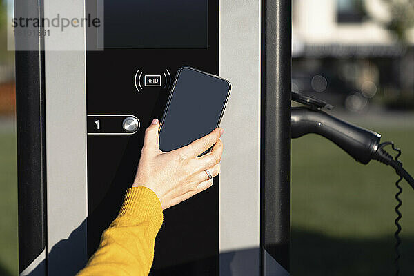 Hand einer Frau  die an der Fahrzeugladestation per Smartphone bezahlt