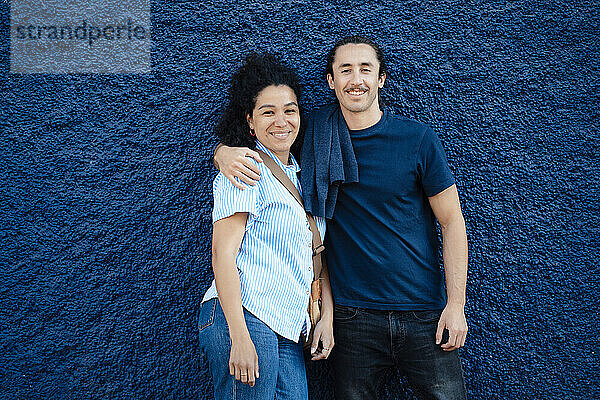 Lächelnder Freund mit dem Arm um seine Freundin vor der blauen Wand