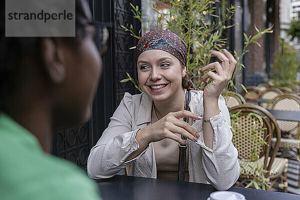 Glückliche Frau mit Kopftuch im Gespräch mit Freundin im Straßencafé