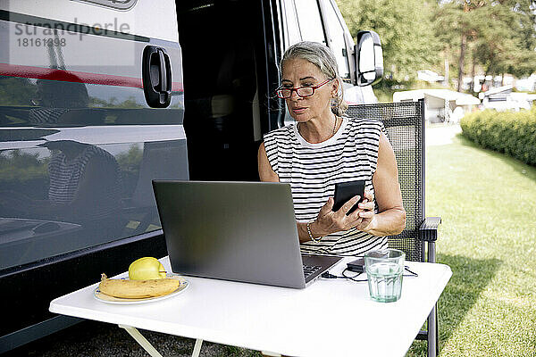 Ältere Frau mit Smartphone und Laptop sitzt am Wohnmobil