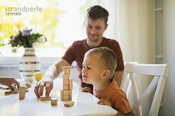 Netter Junge schaut seinem Vater dabei zu  wie er Bauklötze auf dem Esstisch stapelt