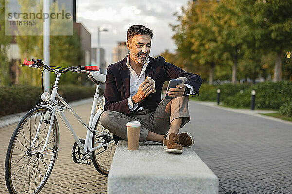 Lächelnder Geschäftsmann benutzt Smartphone und isst mit dem Fahrrad auf der Bank zu Mittag