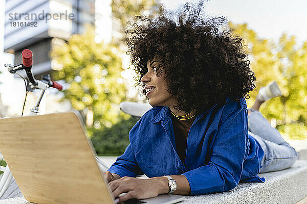 Lächelnde Afro-Frau mit Laptop auf Betonbank liegend