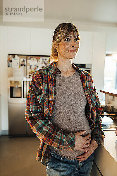 Nachdenkliche schwangere Frau mit den Händen auf dem Bauch steht zu Hause