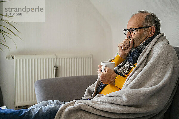 Mann hält Tasse und fühlt sich kalt  während er zu Hause auf dem Sofa sitzt