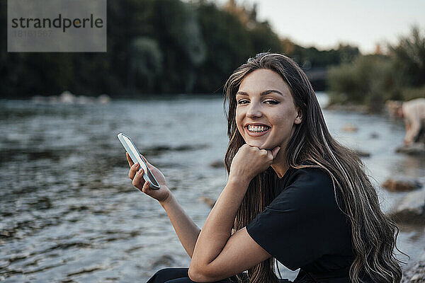 Glückliche junge Frau hält ihr Mobiltelefon und sitzt mit der Hand am Kinn am See